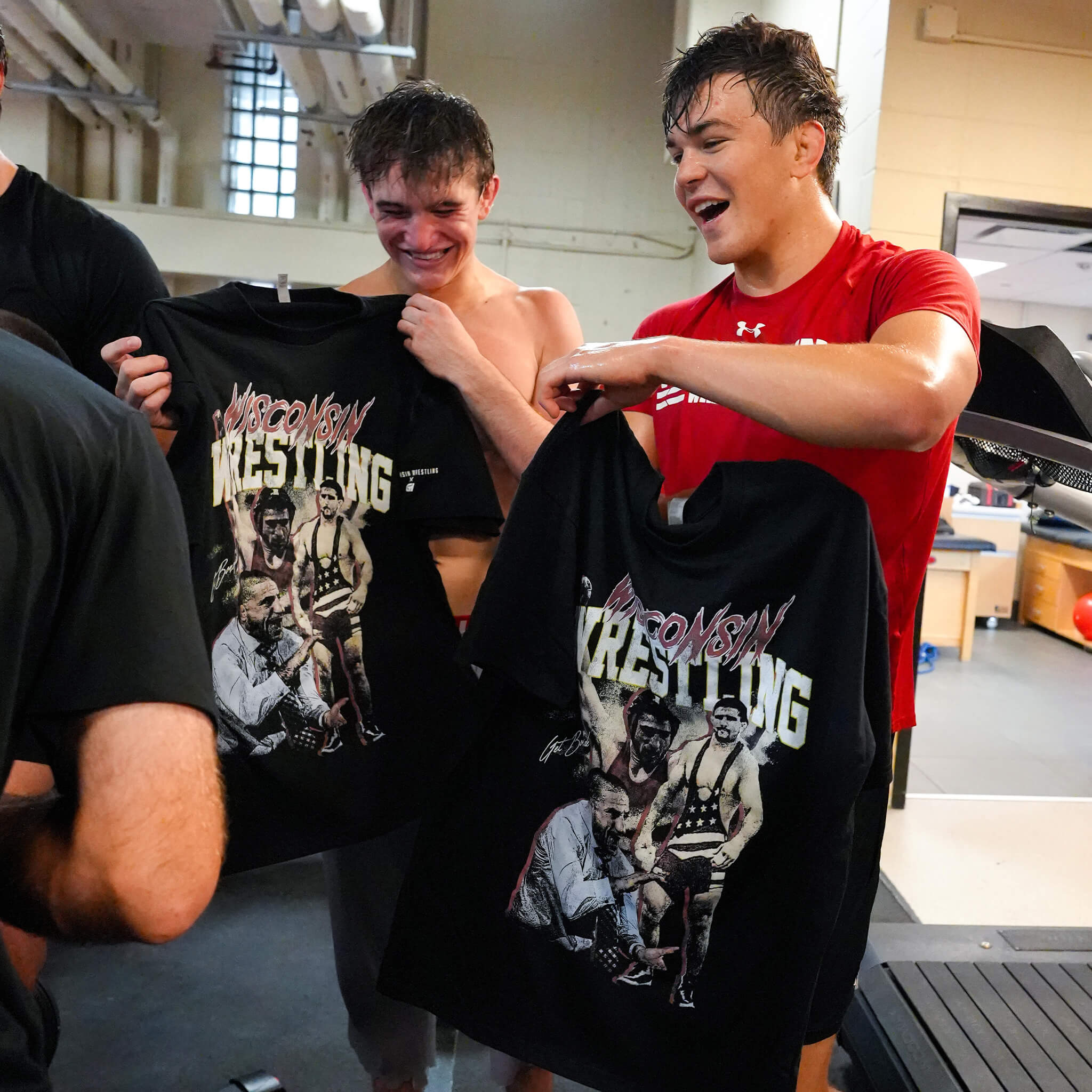 Two smiling wrestlers holding up black CH Gear vintage-style Wisconsin Wrestling t-shirts featuring custom graphic prints in a locker room.