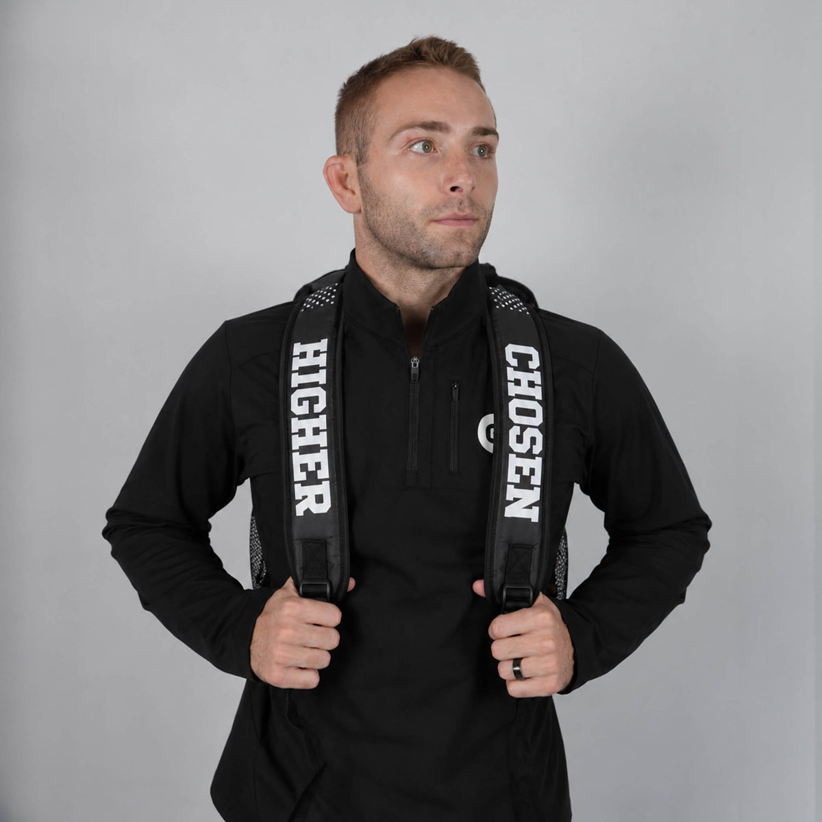 Cayden Henschel wearing a black long-sleeve quarter-zip pullover and a black backpack with white text reading 'HIGHER' and 'CHOSEN' on the straps, posed against a solid gray studio background.