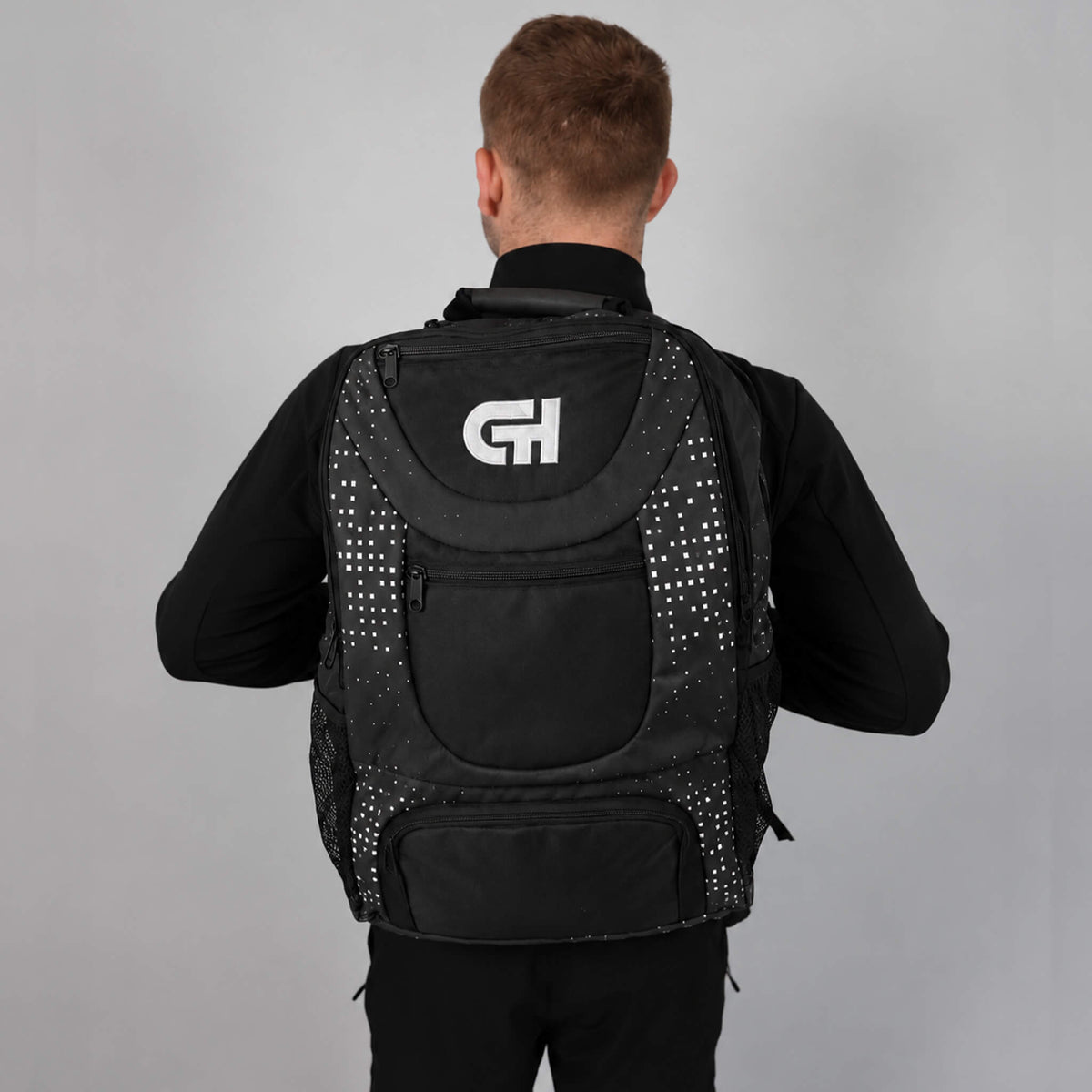 Cayden Henschel viewed from behind wearing a black athletic backpack with white dotted side panels and a white stylized 'CH' logo on the back, set against a plain gray studio background.