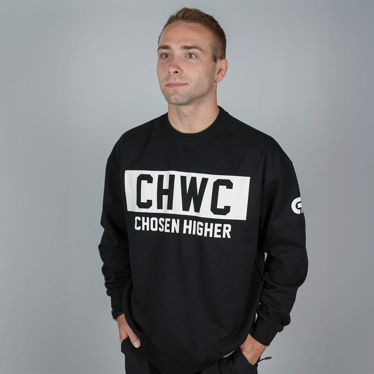 A man wearing a black long-sleeve crewneck sweatshirt with a bold white chest graphic that reads 'CHWC CHOSEN HIGHER' on a plain gray studio background.