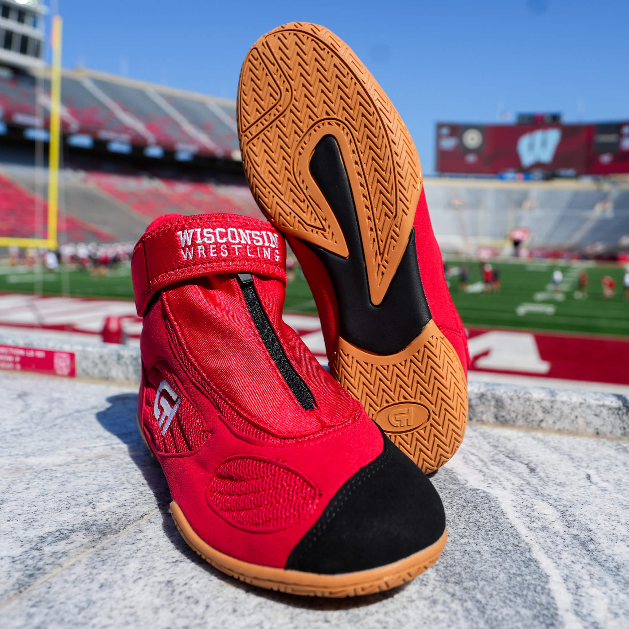 Close-up of red CH Gear Wisconsin Wrestling shoes with gum soles, showing the strap, zipper detail, and tread pattern, set against a blurred football stadium background.