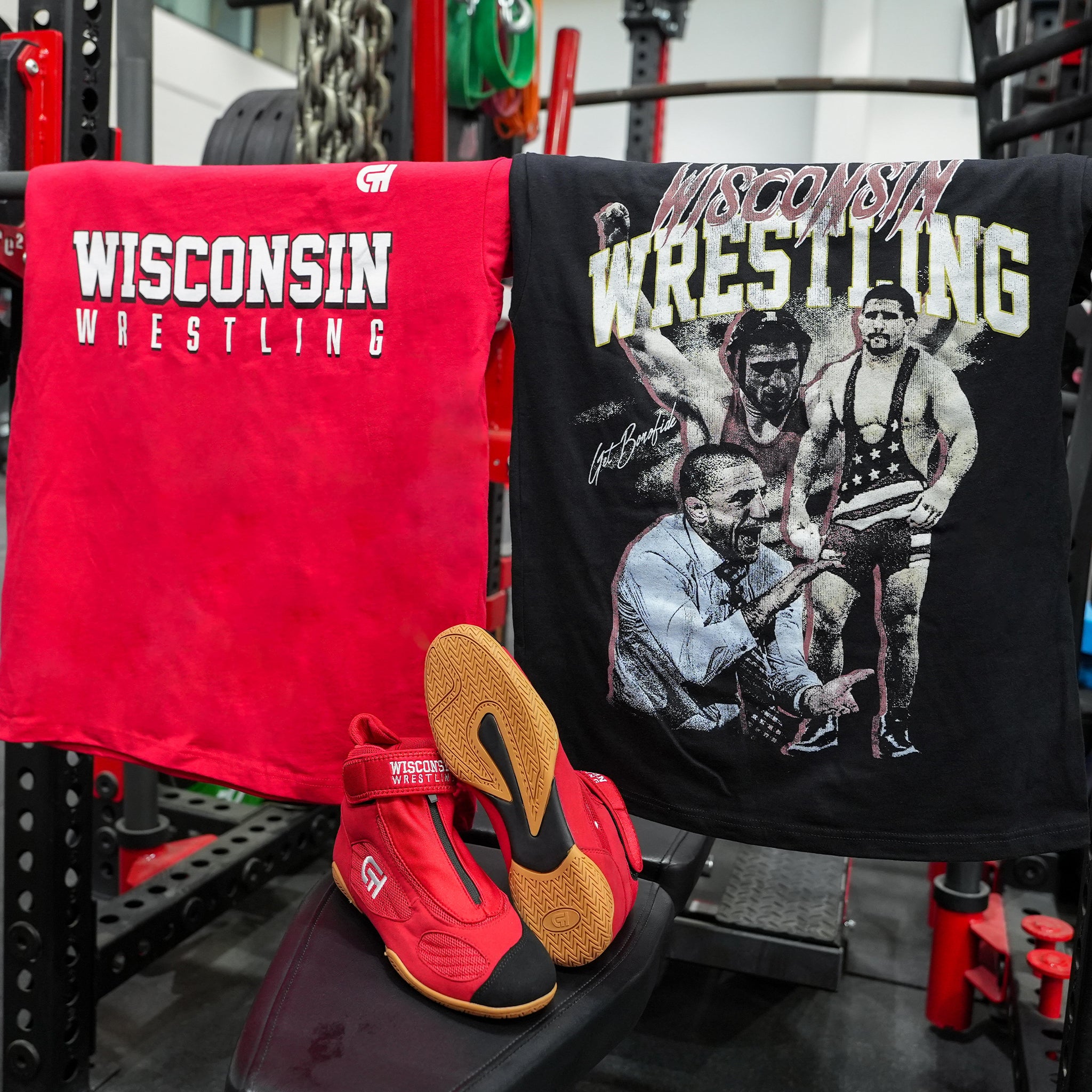 CH Gear red and black Wisconsin Wrestling t-shirts draped over a squat rack in a weight room with red high-top wrestling shoes resting below.