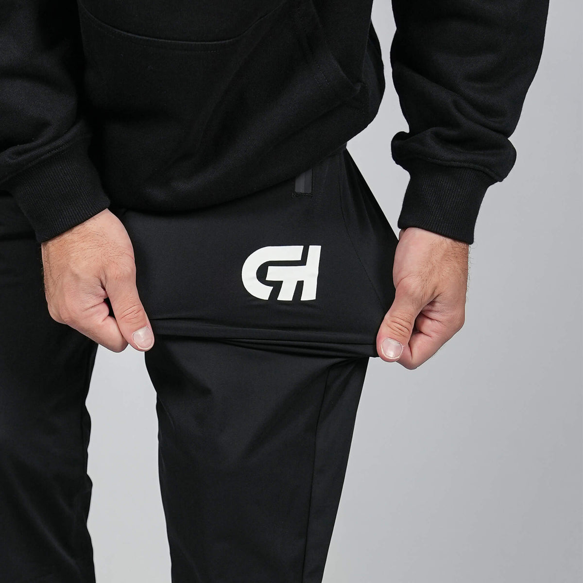 A close-up of a person wearing black sweatpants and a black hoodie, pulling the fabric of the pants to show stretch. The sweatpants feature a white stylized 'CH' logo on the left thigh, set against a plain gray studio background.